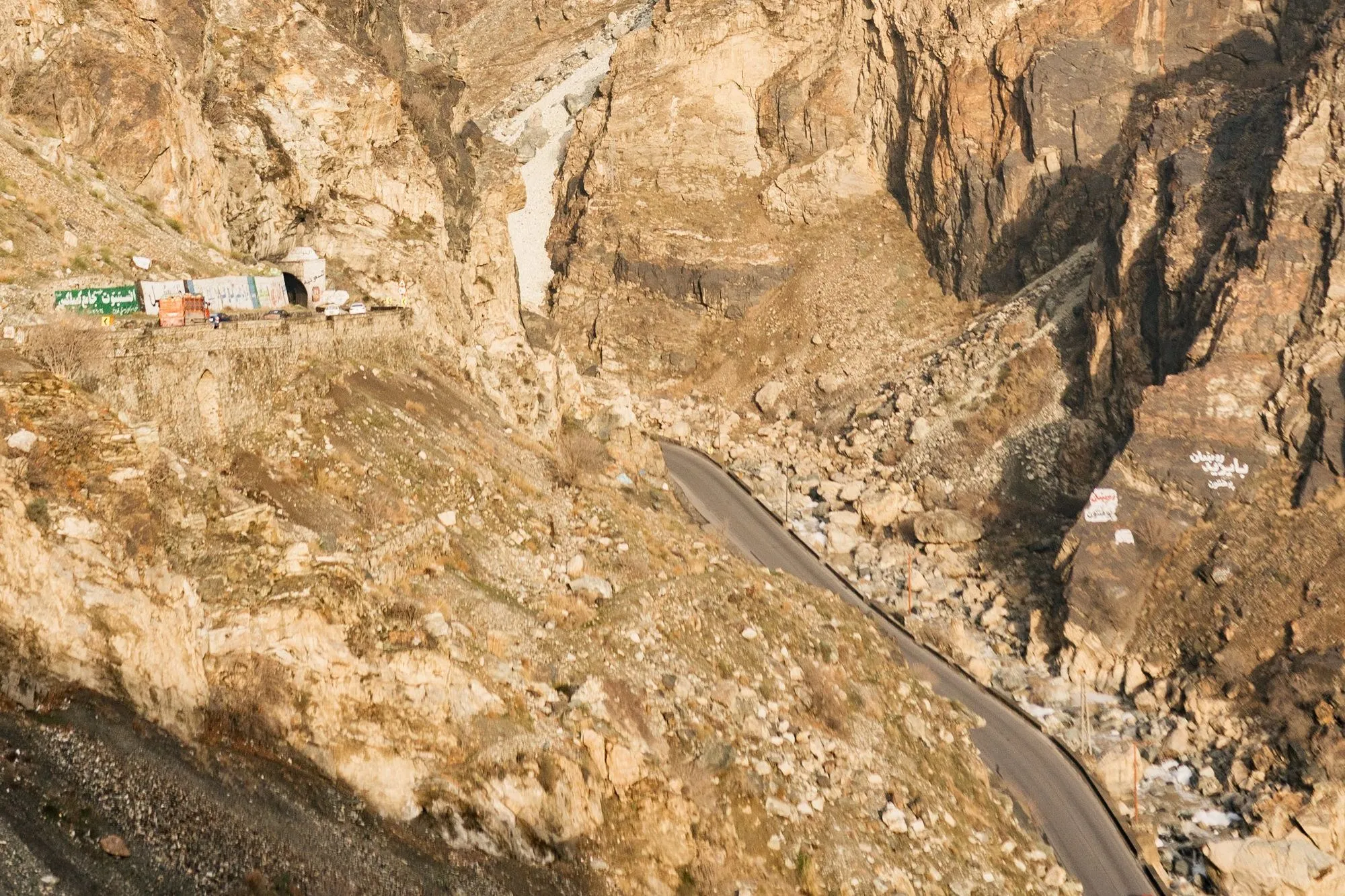 Eine Straße windet sich durch eine steile Felsschlucht, oben am Hang stehen Lastwagen vor einem Tunnel